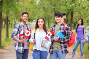 Four students stand outdoors on a green walkway holding Canadian flags and books.