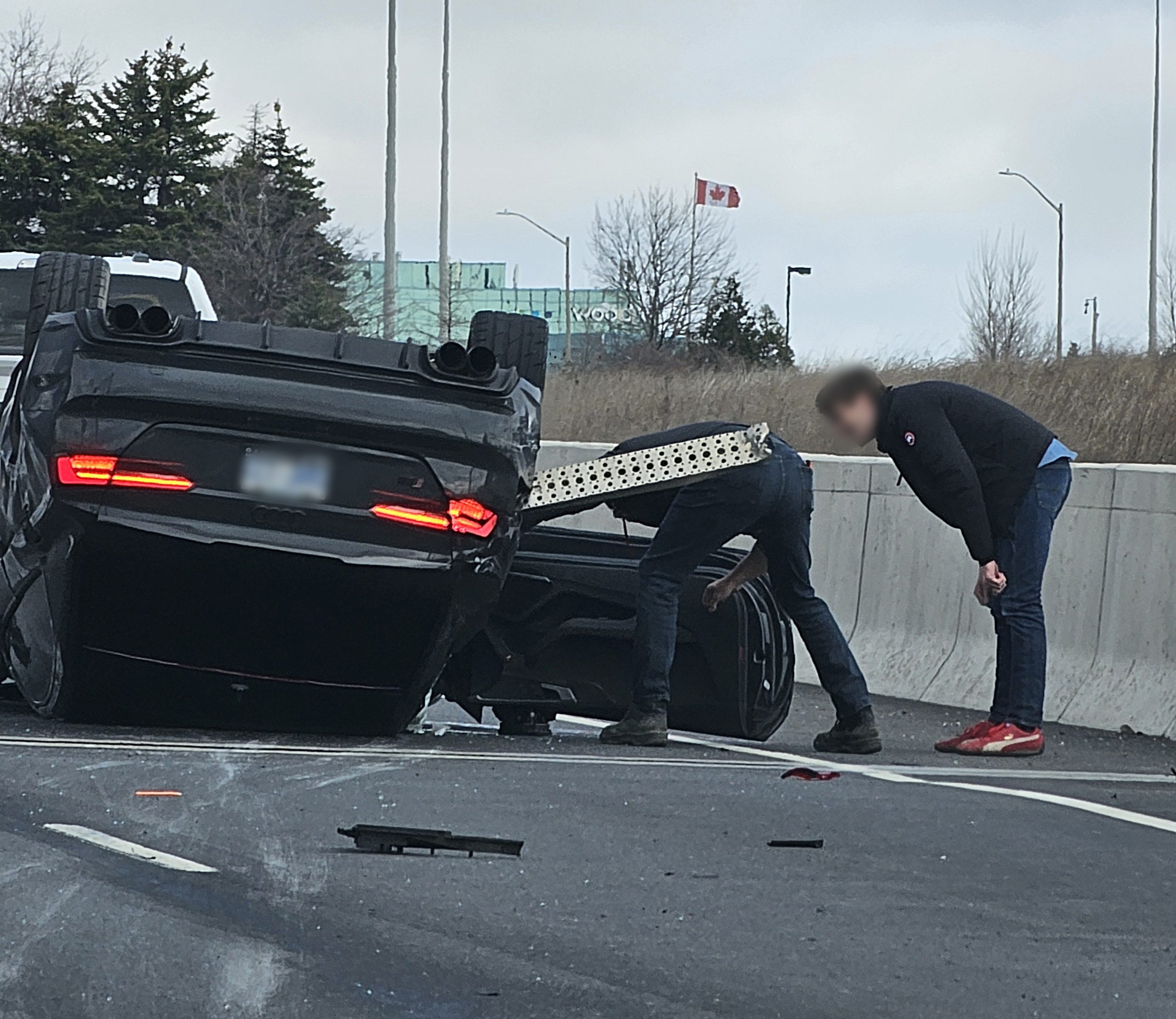2 people trying to help the driver/people inside. Overturned black Audi car on ON-403 highway in Oakville after a severe accident
