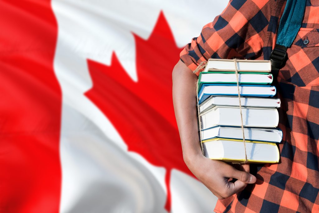 طالب يرتدي ملابس غير رسمية يحمل كومة من الكتب على خلفية العلم الكندي.