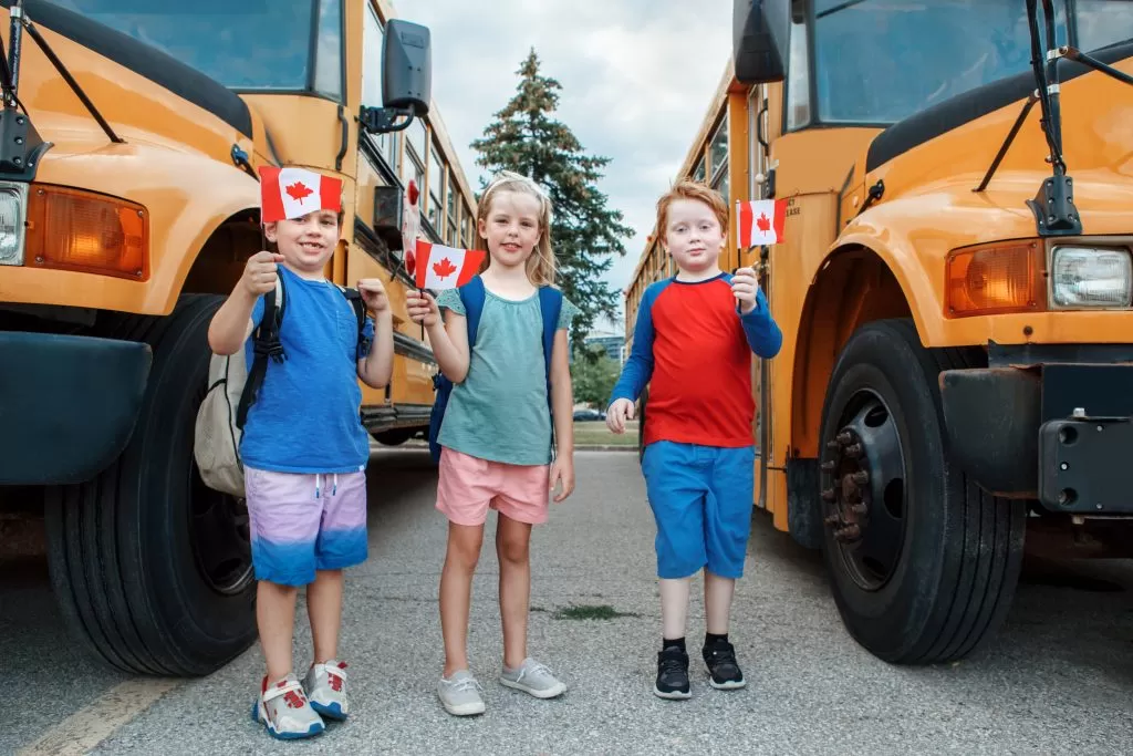 ثلاثة أطفال مبتهجين يحملون الأعلام الكندية أمام الحافلات المدرسية، يحتفلون بيوم كندا و يعبرون عن فخرهم كونهم كندين.