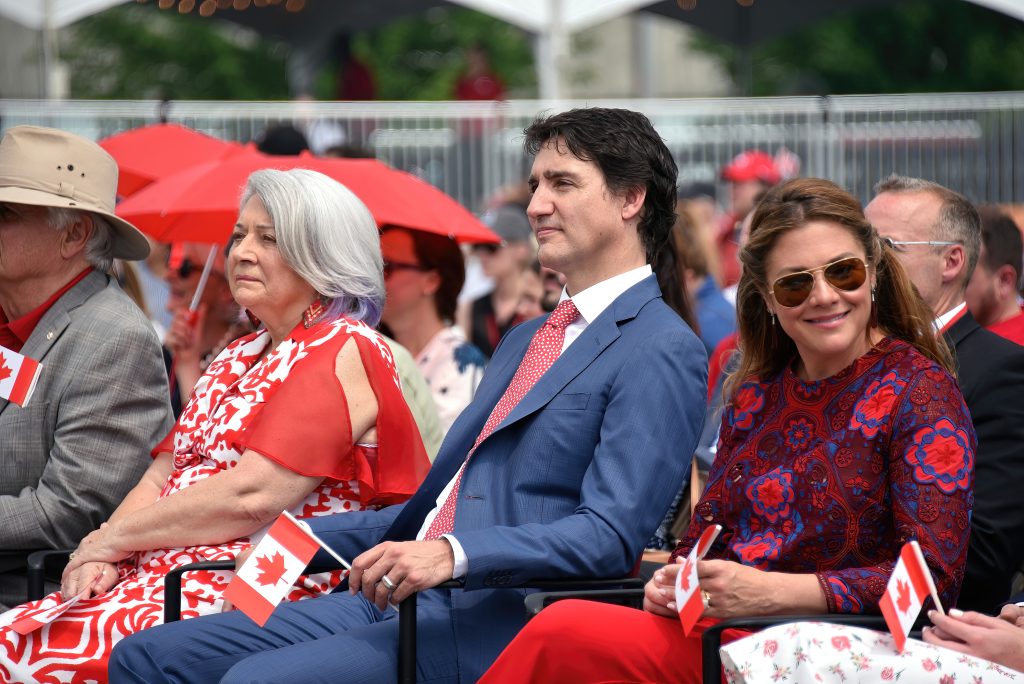 مجموعة من الأشخاص يحضرون حدثًا وطنيًا بقيادة جاستن ترودو الذي يرتدي بدلة مع ربطة عنق حمراء ماسكاً بيده علم كندا صغير.