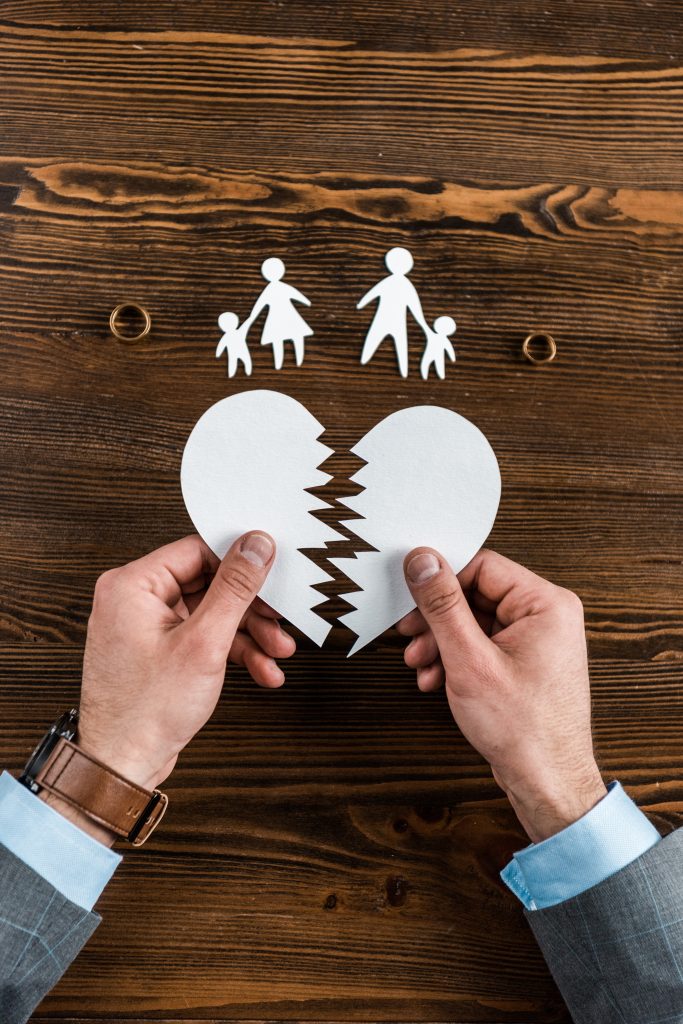 زوج يحمل نصفي قلب ورقي ممزق. تظهر الصورة الخلفية لعائلة مقسمة إلى قسمين على سطح خشبي. يرمز هذا المشهد إلى انفصال الأسرة أو الطلاق.