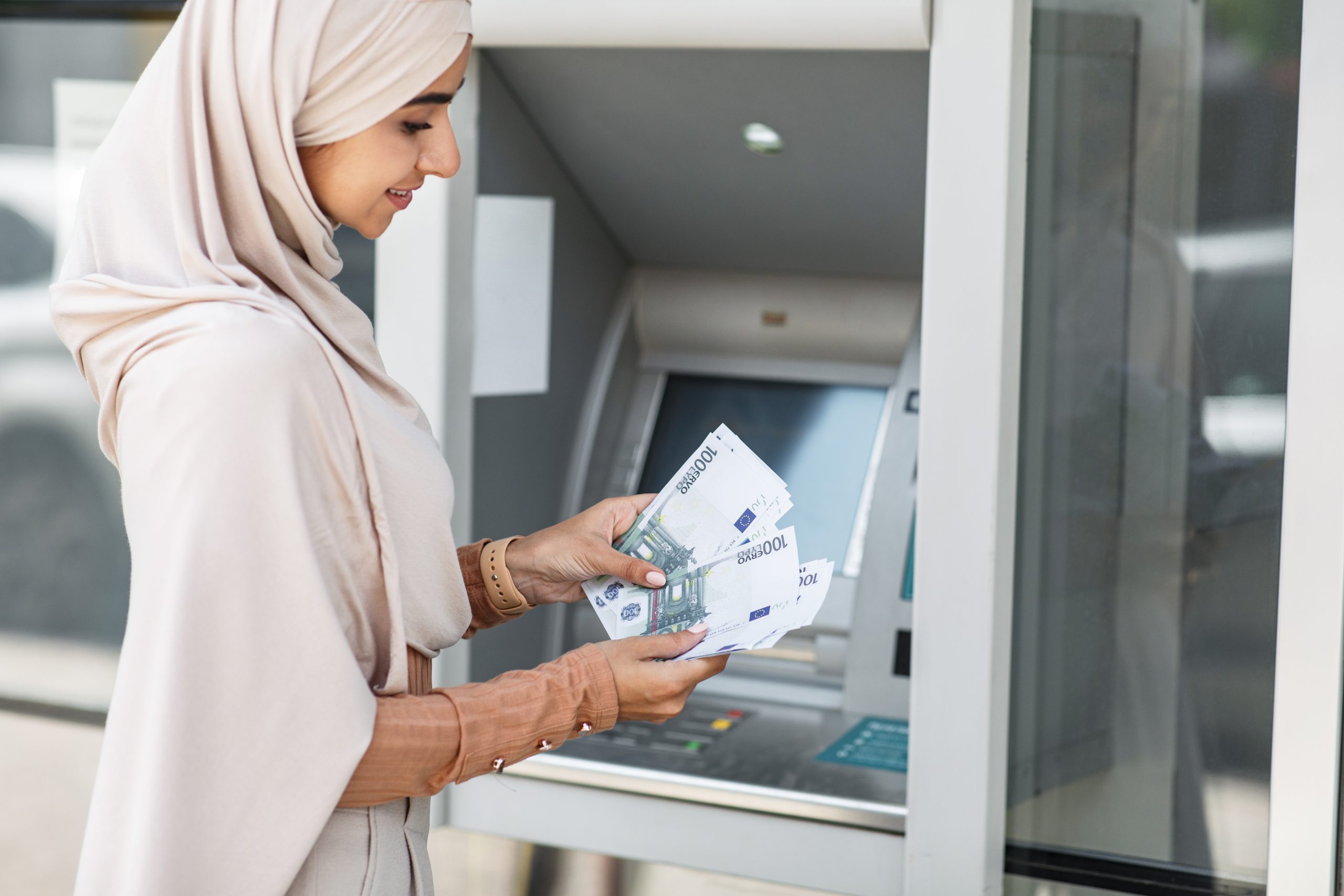 Glad pretty millennial islamic female in hijab counting many euros near atm outdoor