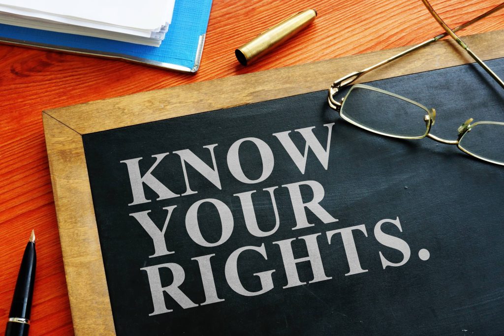سبورة سوداء مكتوب عليها عبارة"اعرف حقوقك" مطبوعة باللون الأبيض، مصحوبة بنظارات ورصاصة ودفتر ملاحظات على مكتب خشبي.