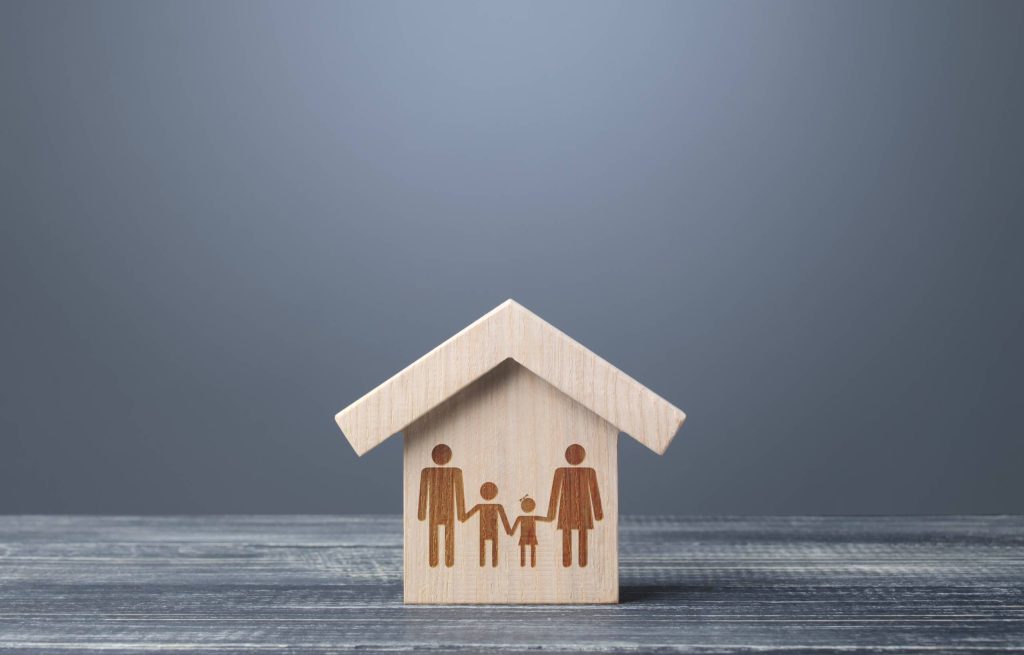 نموذج خشبي لمنزل بداخله أشكال منحوتة لعائلة (شخصين بالغين وطفلين)، على خلفية رمادية.
