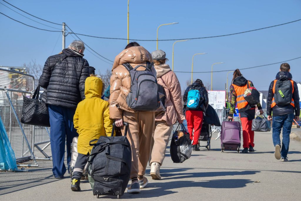 أشخاص يحملون أمتعة يسيرون في أحد الشوارع، يُنظر إليهم من الخلف، مما يشير إلى الهجرة أو البحث عن ملجأ في كندا في يوم مشمس.