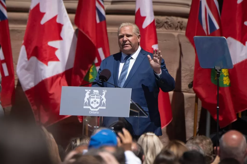 دوغ فورد يتحدث على منصة تحمل شعار أونتاريو، وتحيط به الأعلام الكندية، ويخاطب حشدًا من الناس وضح النهار.