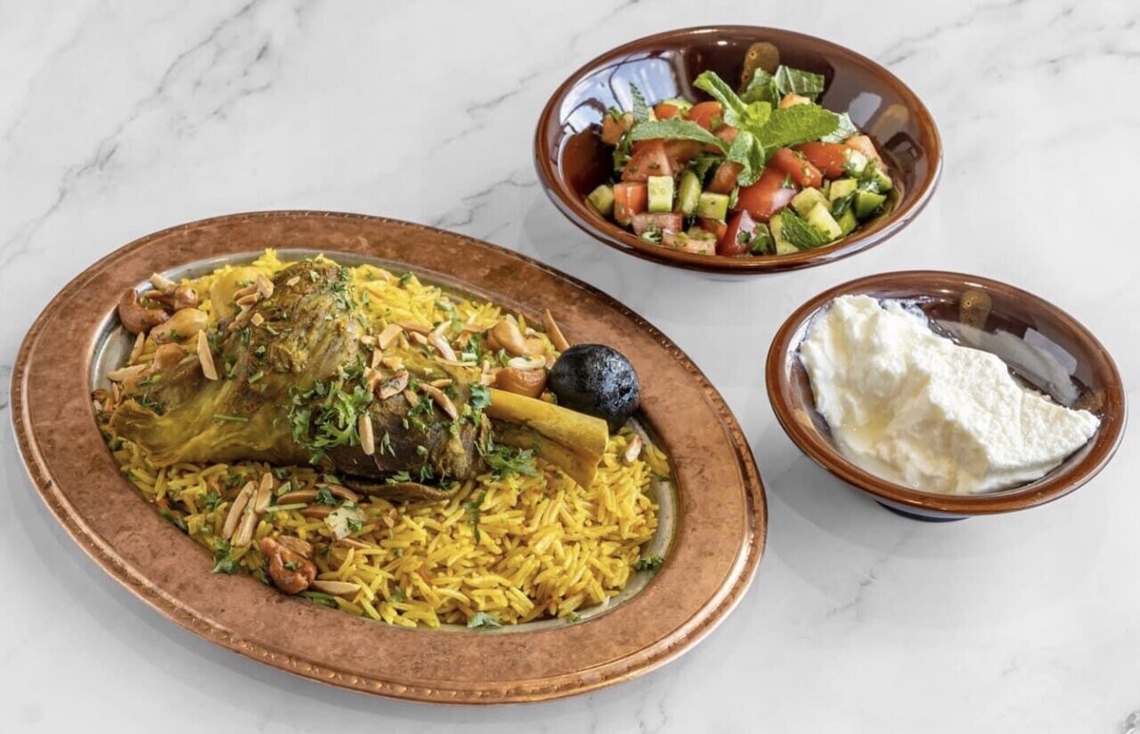 A plate containing yellow rice with a large piece of meat decorated with nuts, a plate of salad, and a plate of white sauce.