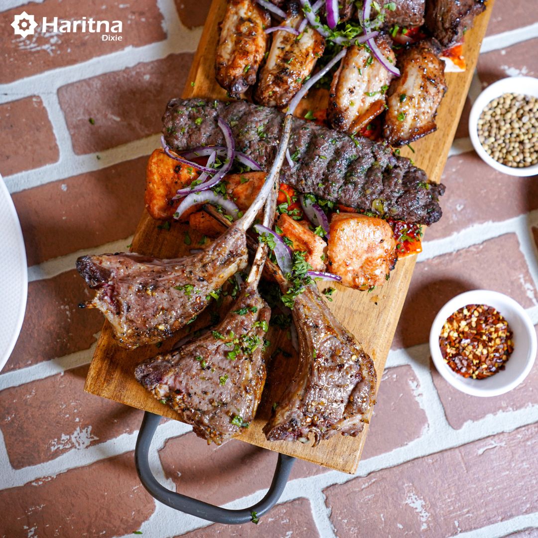 A wooden board filled with different types of grilled meats, including lamb, sausages, and meat skewers, decorated with herbs and onions.