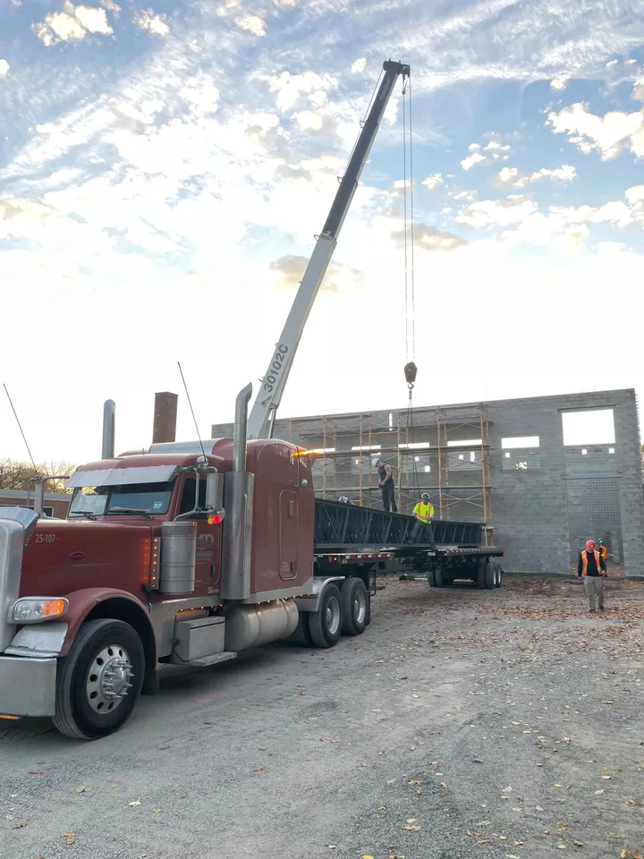 A red semi-trailer truck is parked at a construction site while a crane lifts a large piece of steel with several workers in safety vests.
