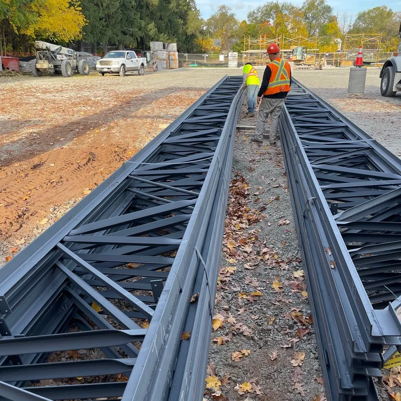 Construction site showing two workers wearing safety vests and helmets walking along a large metal structure that appears to be a steel beam.