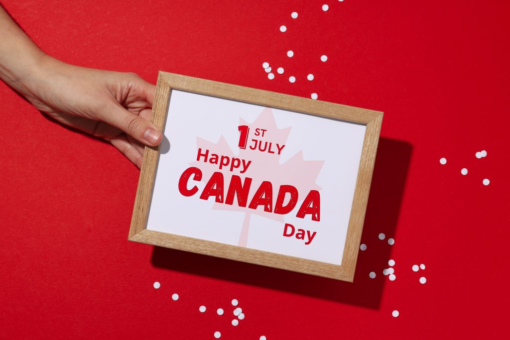يد شخص تمسك بإطار صورة خشبي على خلفية حمراء زاهية. داخل الإطار، توجد رسالة احتفالية باللون الأحمر نصها '1st JULY Happy CANADA Day' (الأول من يوليو، يوم كندا سعيد)، مع صورة باهتة لورقة القيقب في الخلفية. تنتشر قصاصات ورق بيضاء دائرية حول الإطار.