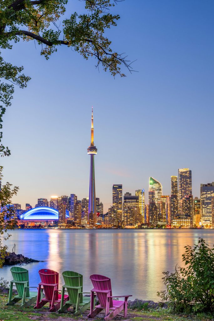 مدينة تورونتو الكندية عند الغروب، يظهر فيها برج سي إن (CN Tower) بشكل بارز مع المباني الشاهقة الأخرى، وتنعكس أضواء المدينة على مياه بحيرة أونتاريو الهادئة.